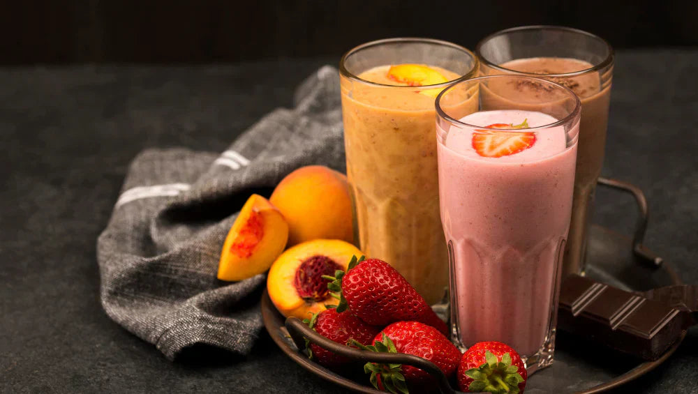 Plusieurs milkshakes protéinés présenté sur une table grises avec des fraises et peches sur un fond noir