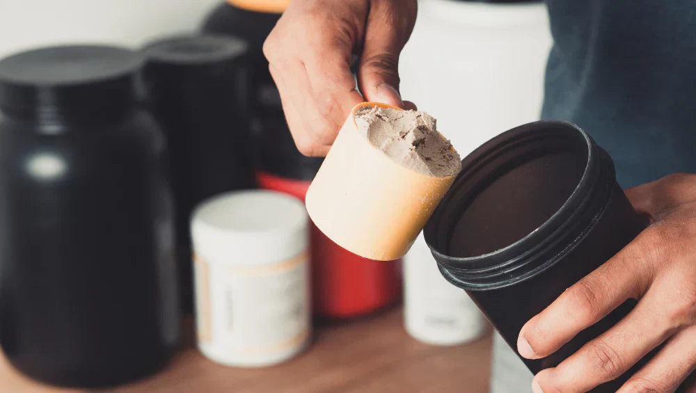 Un homme en train de mettre une scoop de proteine dans un shaker noir avec dansd le fond plusieurs pot de proteine ainsi que d'autres compléments alimentaires