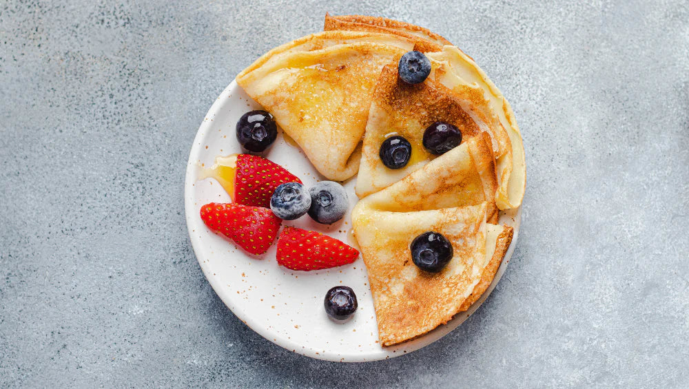 Crêpes protéinées dans une assiette blanche entouré de myrtille et de fraises posé sur un fond gris