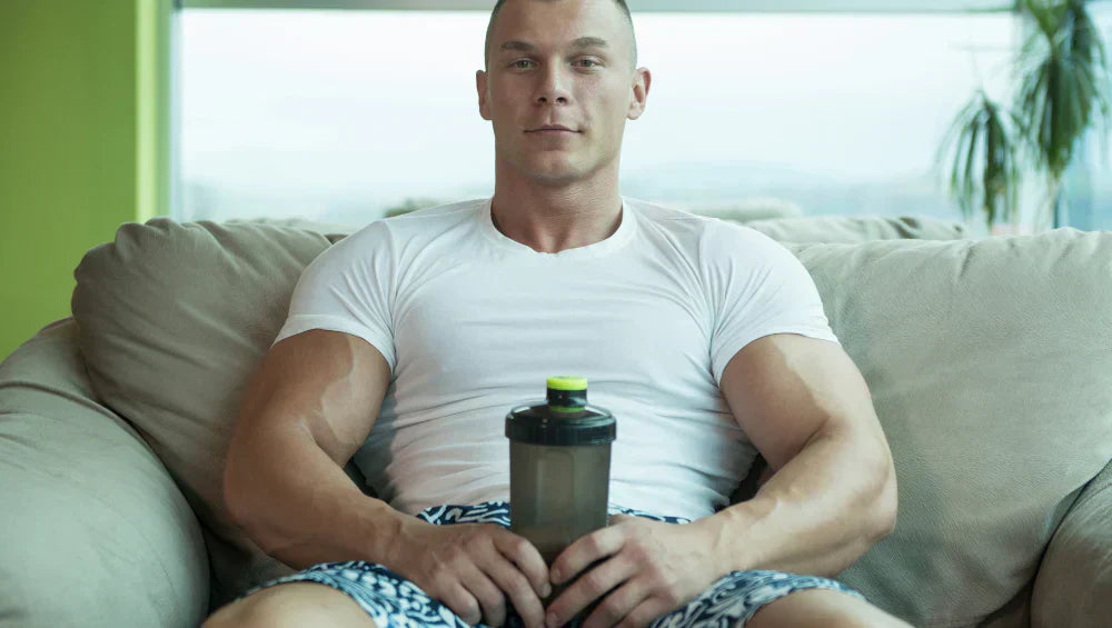 Homme musclé dans son canapé en train de tenir un shaker de protéine dans ses main