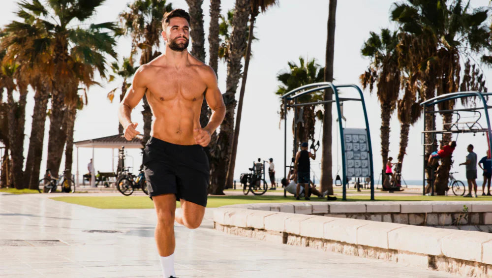 Homme musclé en train de courir en short noir et torse nu sur les bord d'une plage avec des palmier en arrière plan